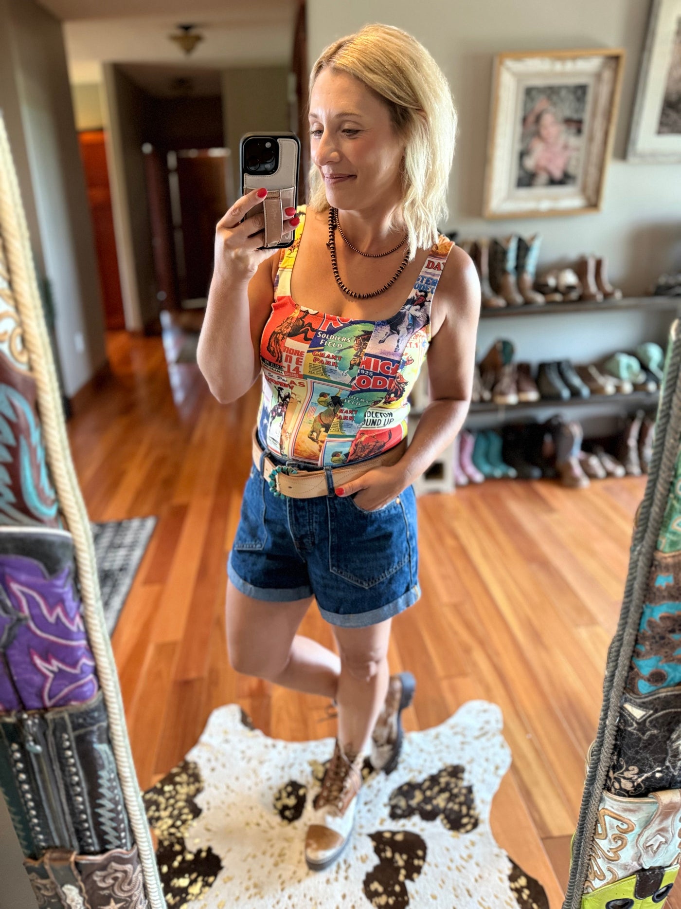 Woman taking a mirror selfie wearing a colorful rodeo magazine style tank top and Western denim shorts accented by a turquoise belt buckle.