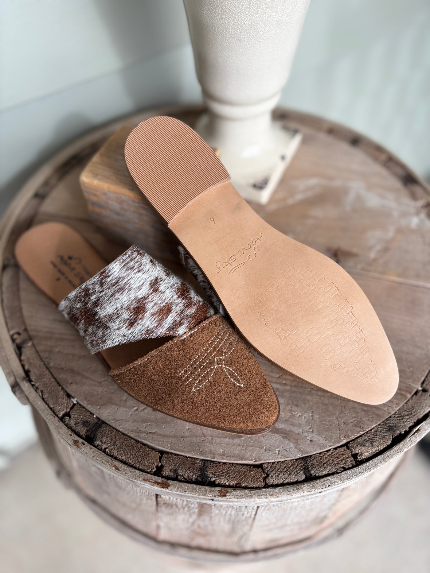 Pair of brown leather mule slides with a tan insole and authentic cowhide on a wooden block.