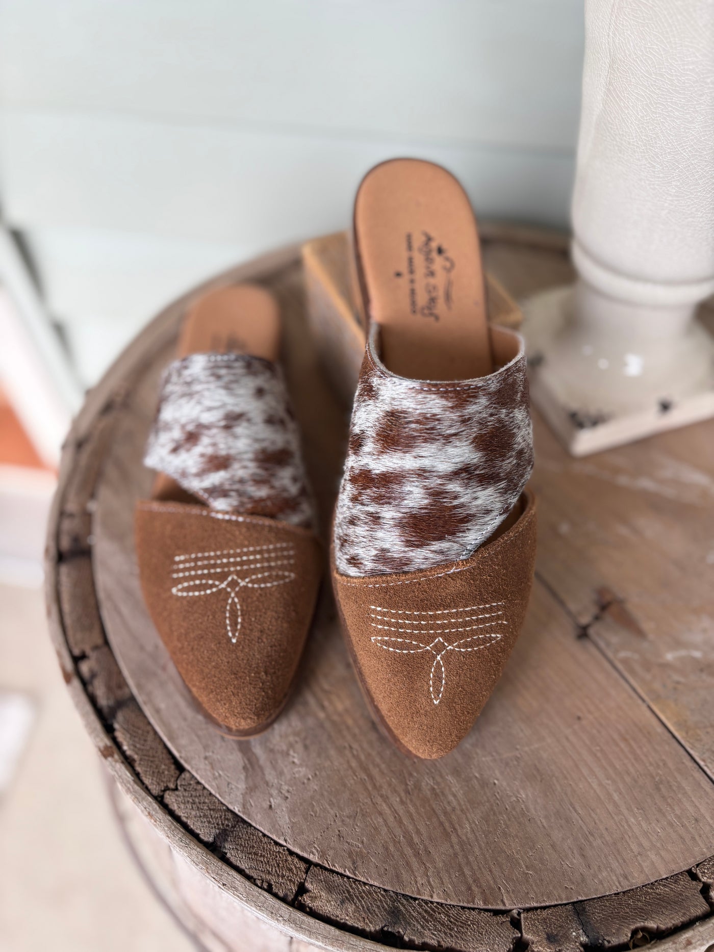 Pair of brown mules with genuine cowhide and suede featuring a bootstitch toe on a wooden surface.
