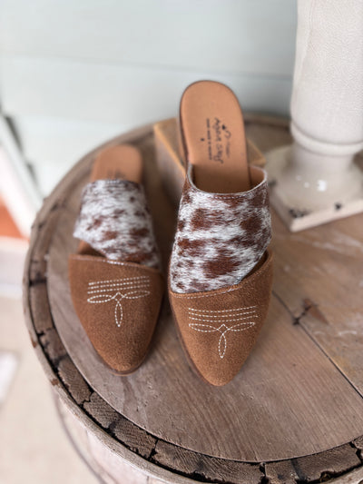 Pair of brown mules with genuine cowhide and suede featuring a bootstitch toe on a wooden surface.