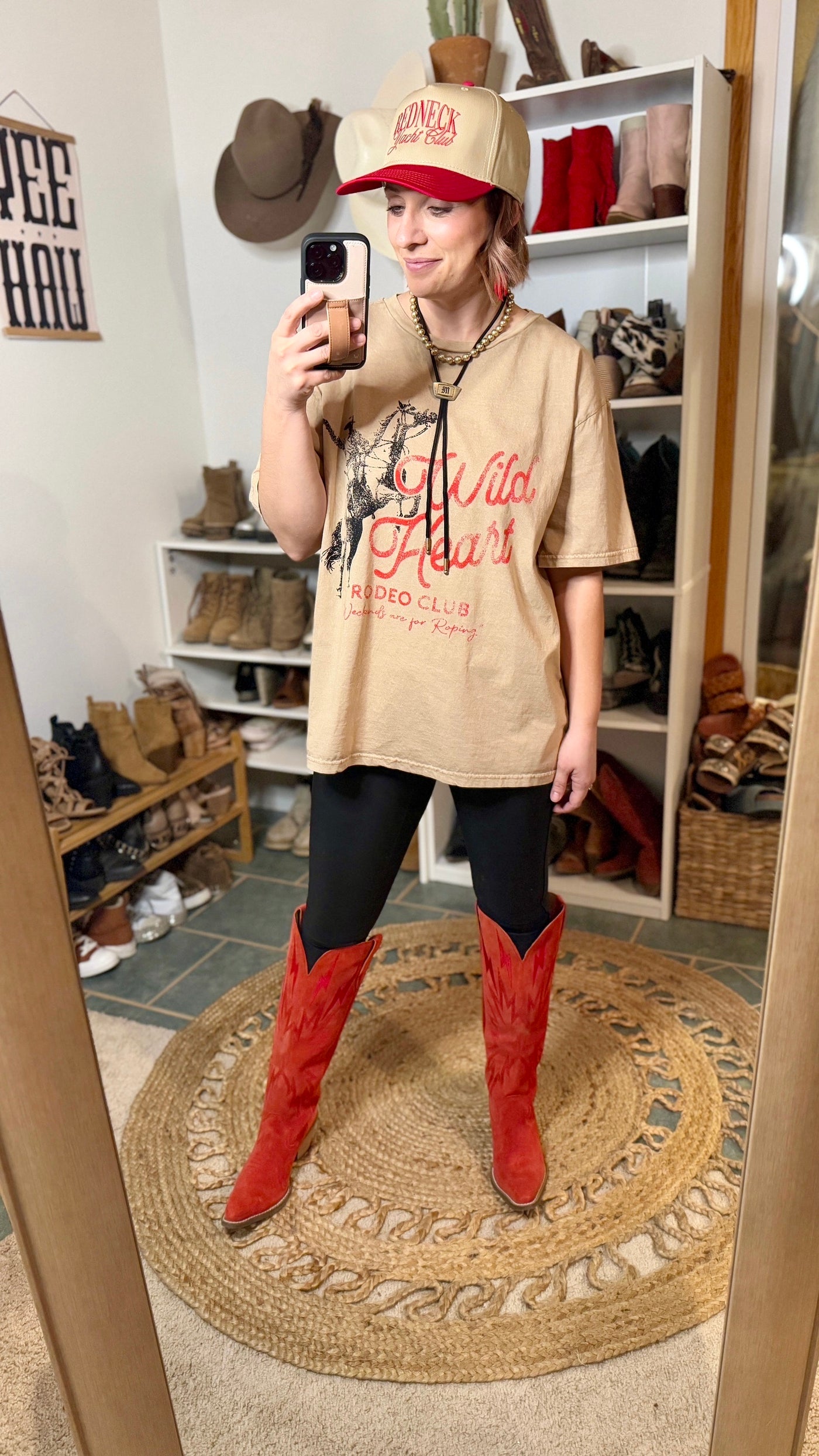 Cowgirl wearing the Bentlee Wild Heart Tee layered over black leggings, featuring an edgy Western aesthetic with finishing touches of a trucker hat and red cowgirl boots.