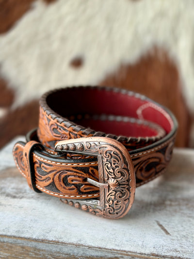 Western-style tooled leather belt with copper-colored engraved buckle, displayed on wooden shelf with cowhide background.