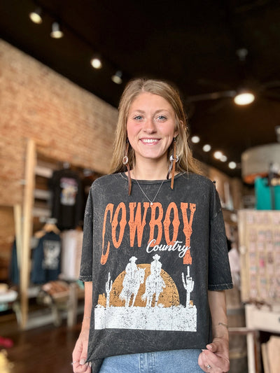 An employee modeling our Frost Cowboy Country Graphic Tee that features the saying "Cowboy Country" and a scene of two cowboys on horseback in the desert which is available at Broker Leather in Waukon, Iowa.