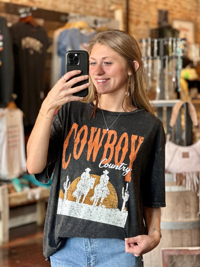 A Broker Leather employee modeling our Frost Cowboy Country Graphic Tee that features the saying "Cowboy Country" and a scene of two cowboys on horseback in the desert.