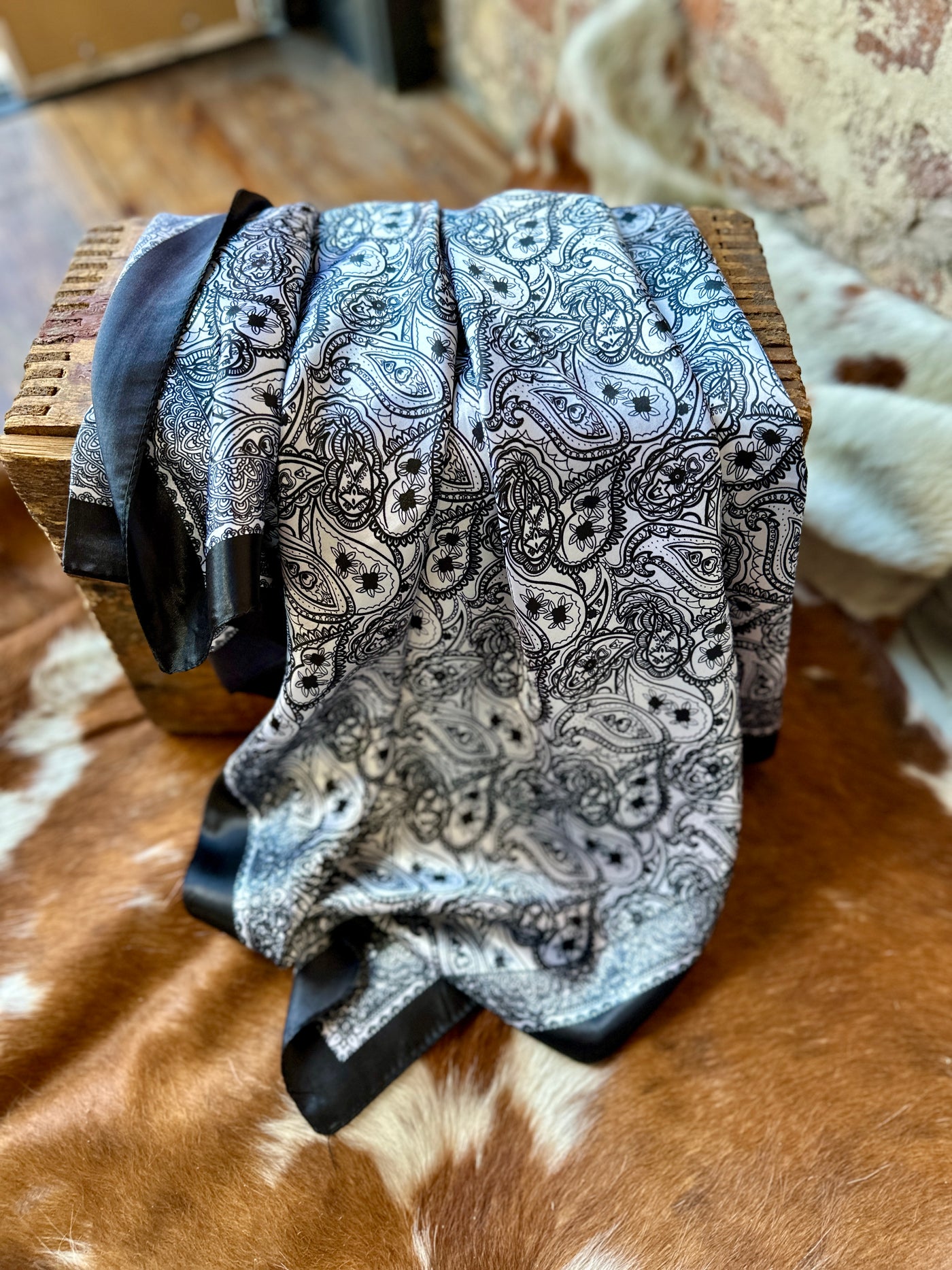 Flatlay photo showcasing our Postal Service Black & White Paisley Wild Rag with a cowhide background.