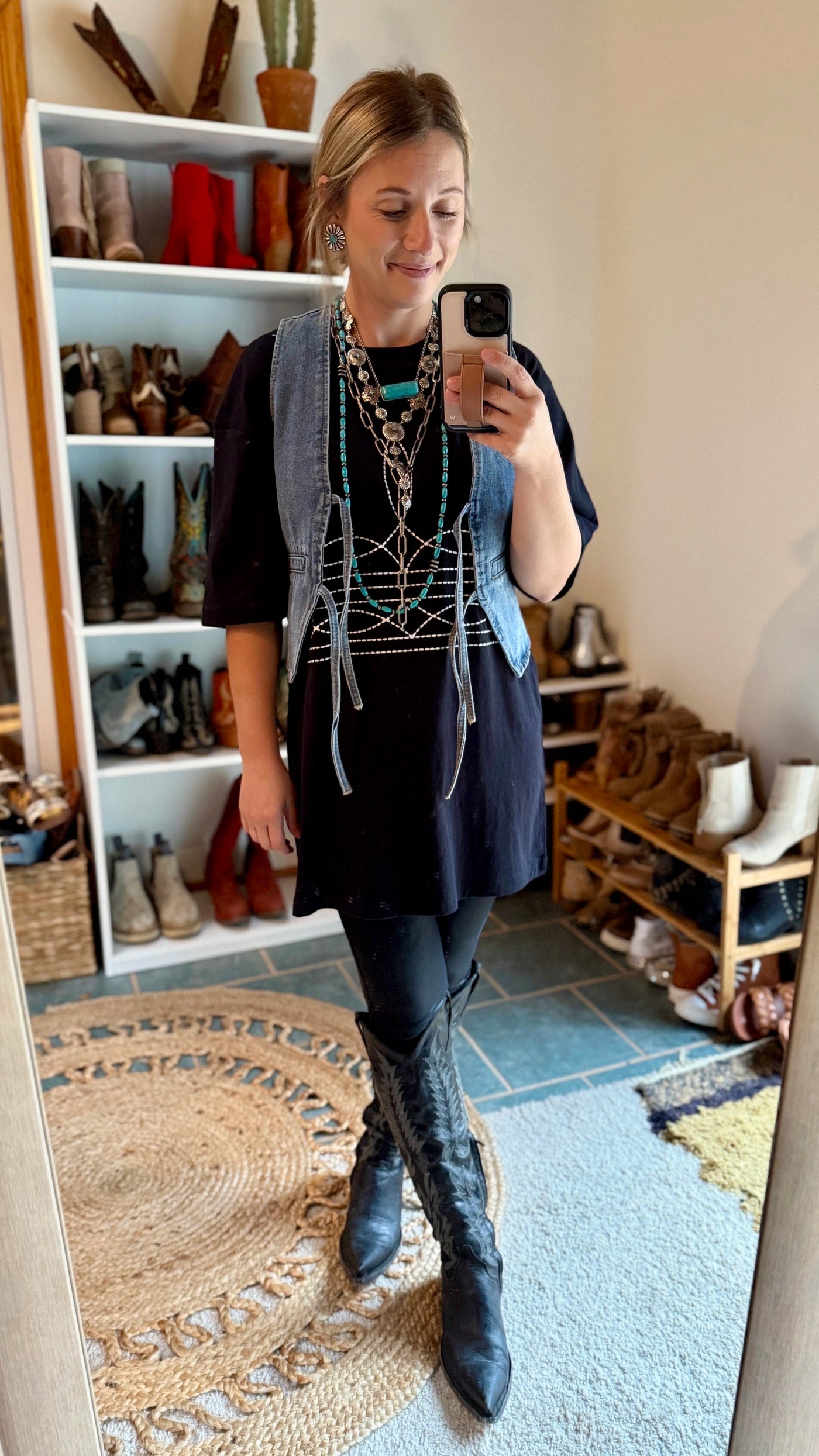 A Broker Leather employee modeling the black Brewer Boot-Stitch Graphic Tee Dress over leggings with boots, statement necklaces, and a denim vest.