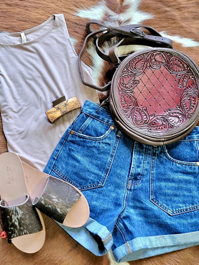 Western outfit inspiration flatlay from Broker Leather featuring denim shorts, a basic tank top, cowhide slides, and a hand-tooled leather canteen style bag.