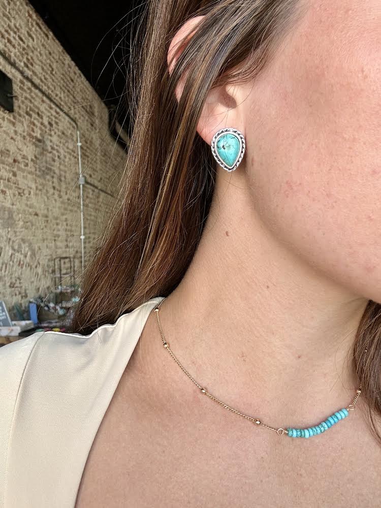 Close-up of teardrop-shaped faux turquoise stud earrings with silver trim modeled by a woman for size reference.