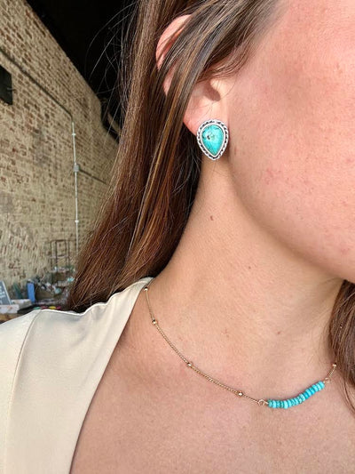 Close-up of teardrop-shaped faux turquoise stud earrings with silver trim modeled by a woman for size reference.