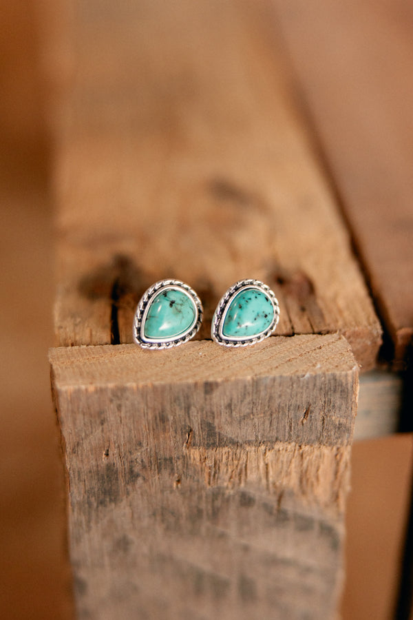 Close-up of teardrop-shaped faux turquoise stud earrings with silver trim on a Western-style background.