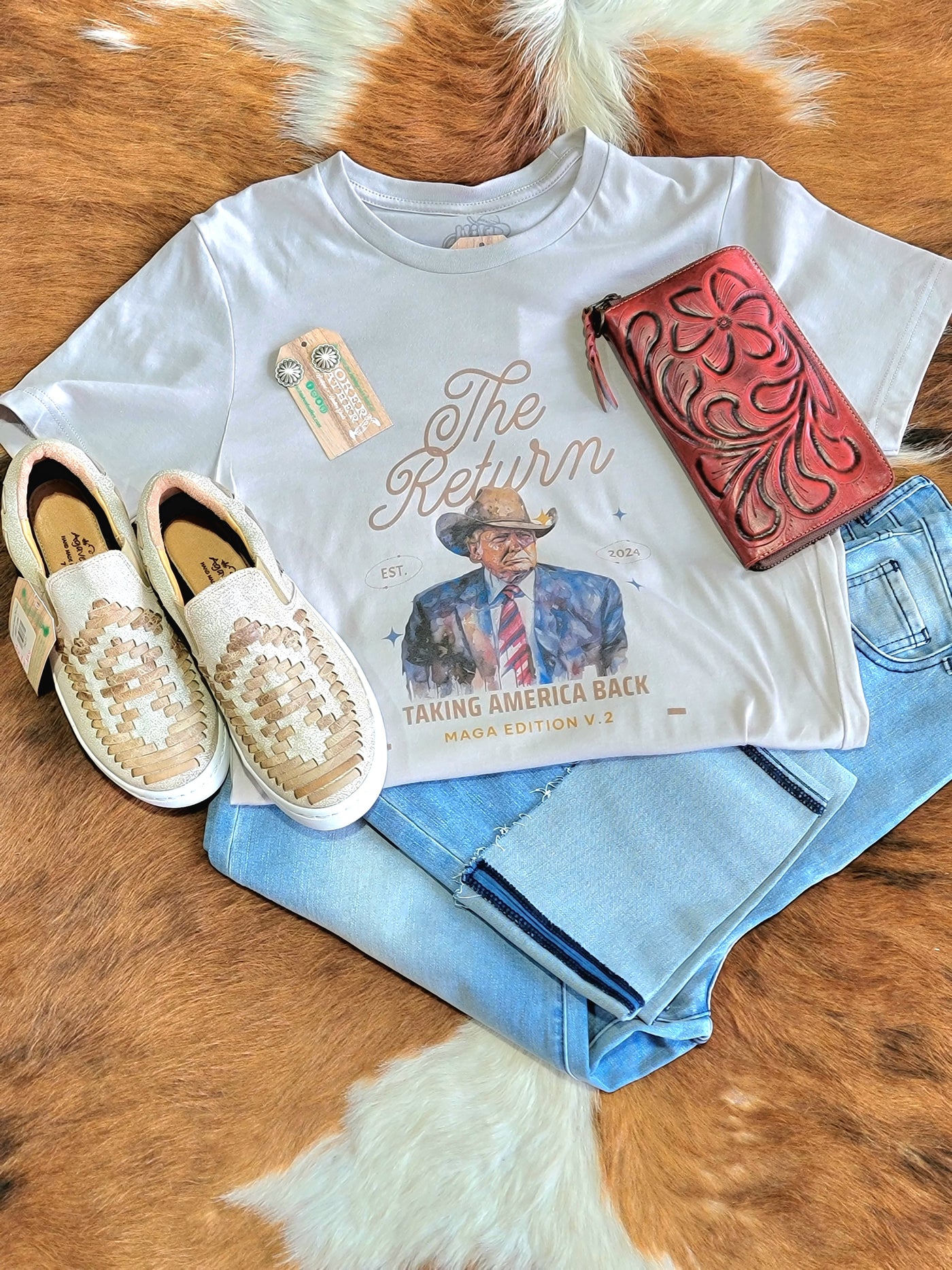 Flatlay photo of the Joram Cowboy Trump Graphic Tee with jeans, leather sneakers, a wallet, and earrings.