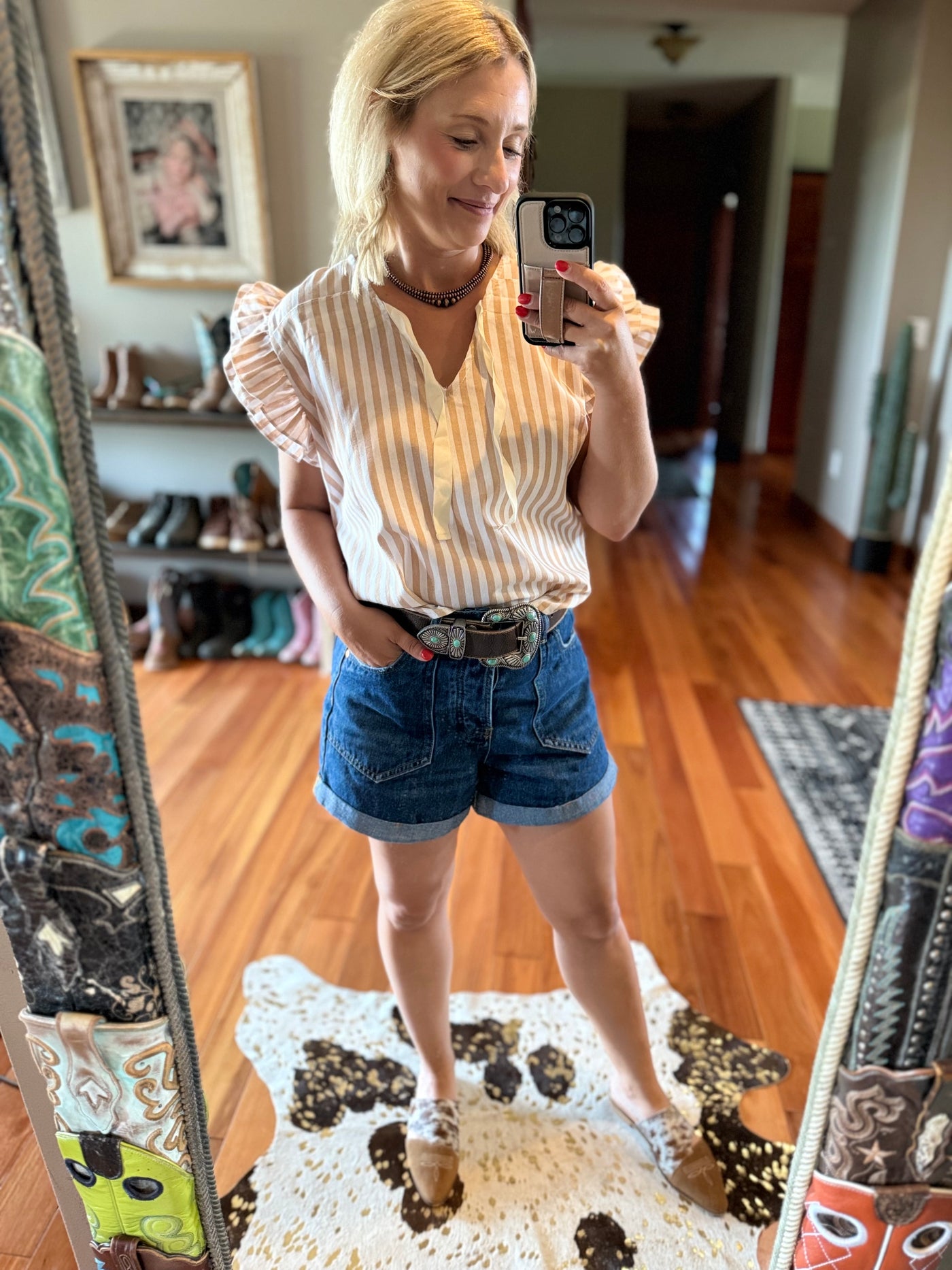 Broker Leather model taking a mirror selfie wearing a striped shirt with ruffled sleeves and a bow tie with a denim shorts in a home setting.