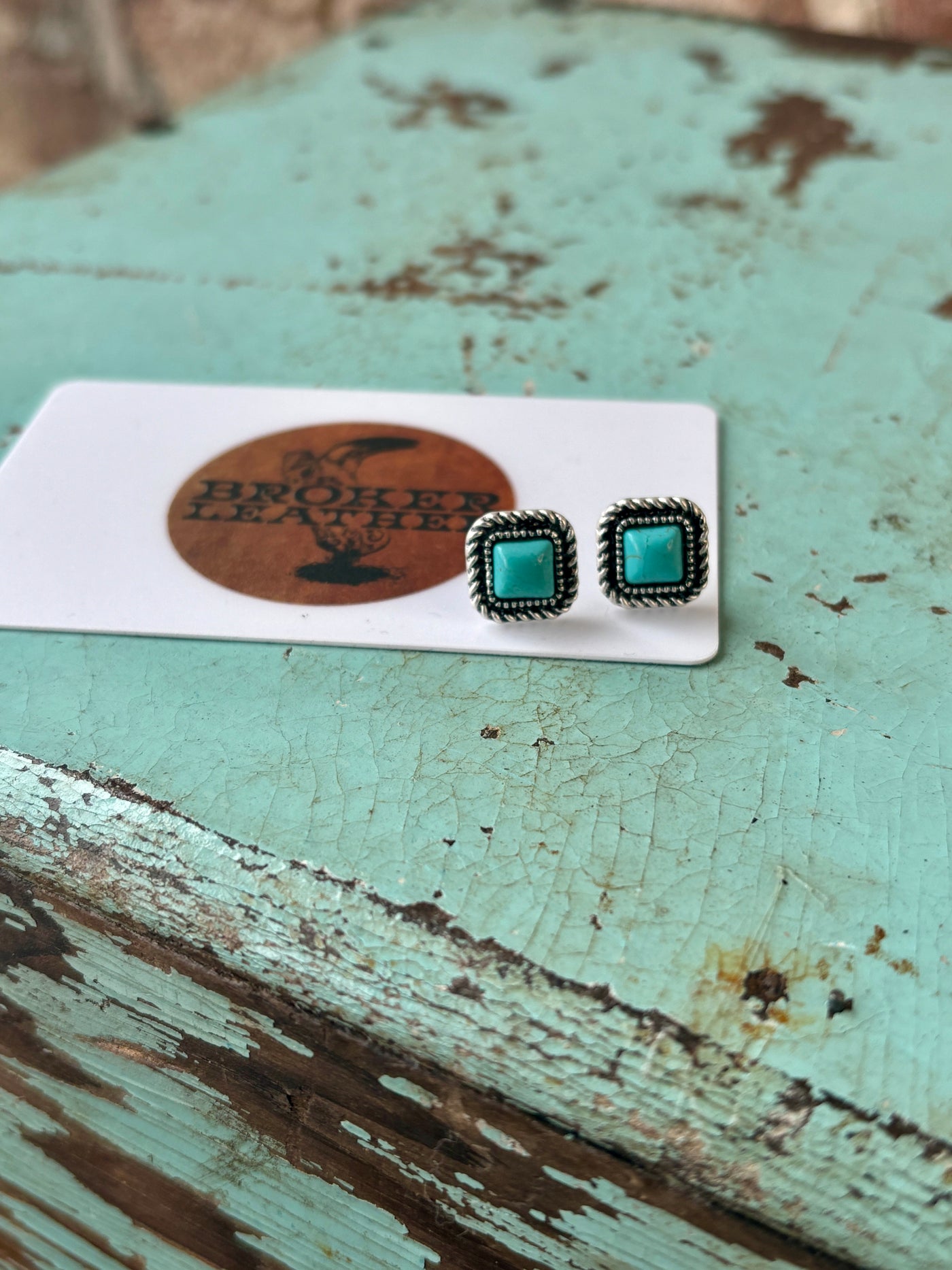 Close-up of faux turquoise stud earrings with intricate western-inspired detailing photographed next to a plastic gift card for size reference.