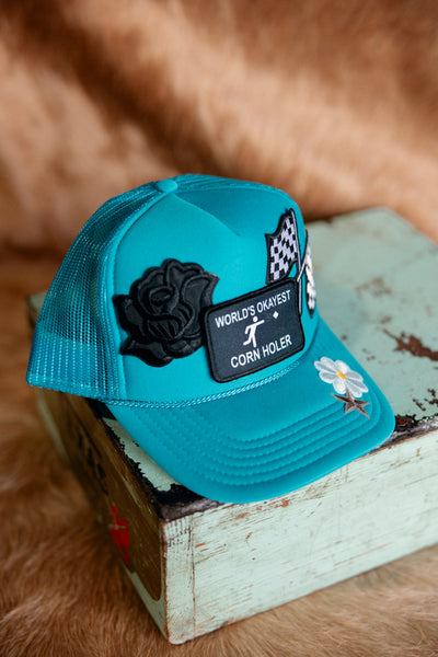 A flatlay photo of a custom World's Okayest Corn Holer Trucker Hat with a cowhide and wood background.